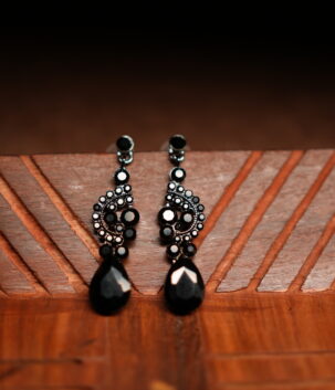 A pair of gunmetal-tone chandelier drop earrings with an ornate black crystal cluster body and a large faceted black teardrop pendant, displayed on a geometric carved wooden surface against a dark background.