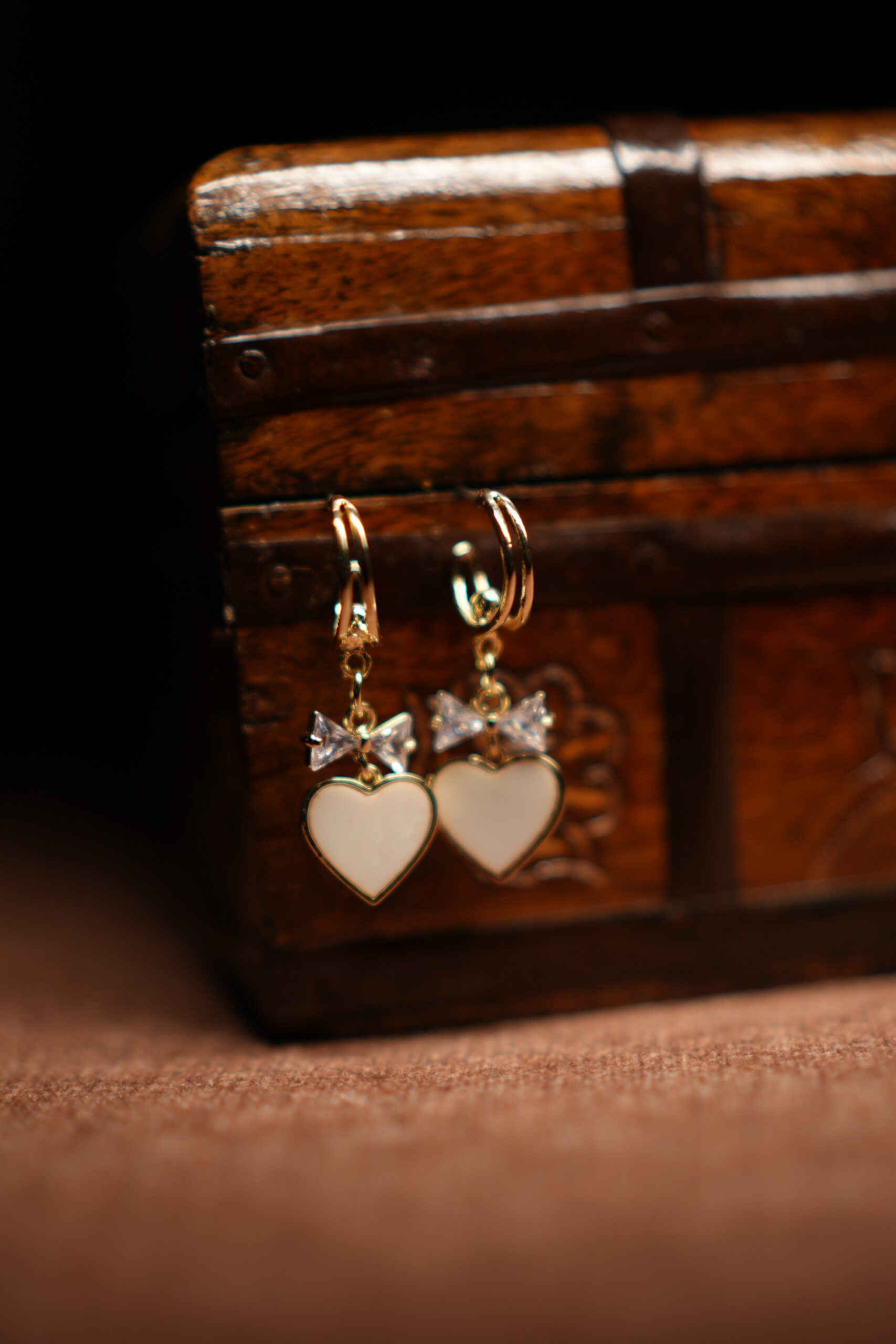 A pair of gold-tone mini hoop earrings with a baguette crystal bow charm and a white enamel heart pendant, displayed hanging from the edge of a vintage carved wooden jewellery chest against a dark background.