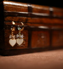 A pair of gold-tone mini hoop earrings with a baguette crystal bow charm and a white enamel heart pendant, displayed hanging from the edge of a vintage carved wooden jewellery chest against a dark background.
