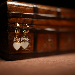 A pair of gold-tone mini hoop earrings with a baguette crystal bow charm and a white enamel heart pendant, displayed hanging from the edge of a vintage carved wooden jewellery chest against a dark background.
