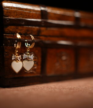A pair of gold-tone mini hoop earrings with a baguette crystal bow charm and a white enamel heart pendant, displayed hanging from the edge of a vintage carved wooden jewellery chest against a dark background.