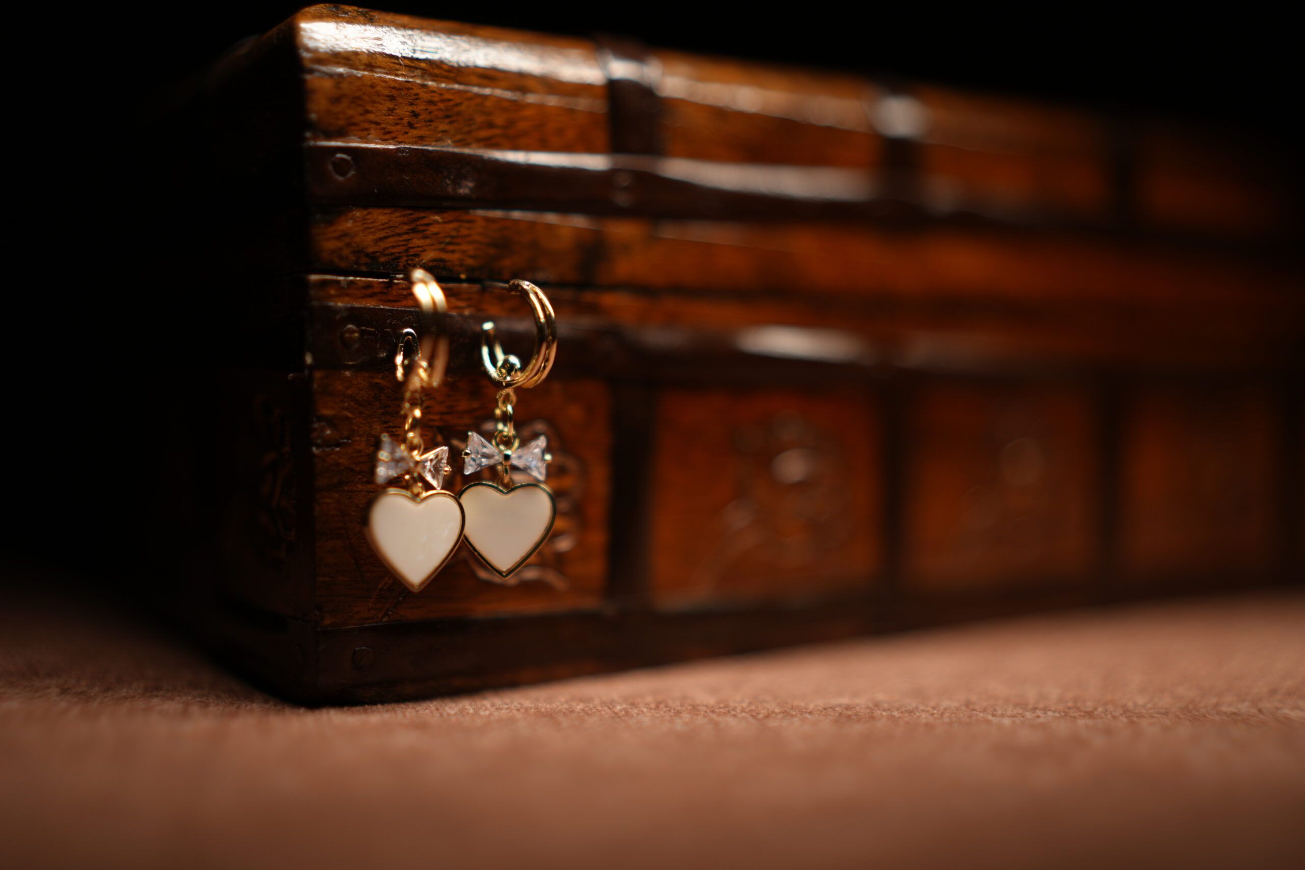 A pair of gold-tone mini hoop earrings with a baguette crystal bow charm and a white enamel heart pendant, displayed hanging from the edge of a vintage carved wooden jewellery chest against a dark background.