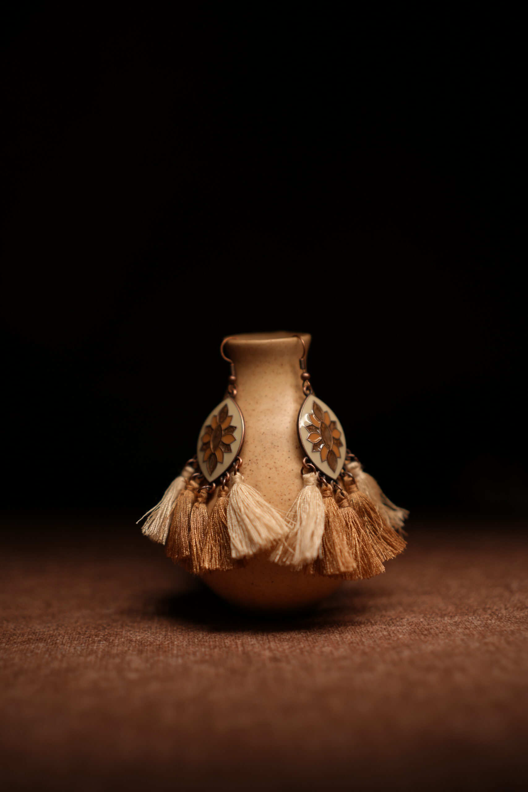 A pair of ivory and gold lotus enamel marquise-shaped drop earrings with five alternating ivory cream and champagne gold thread tassels, displayed on a speckled beige ceramic vase against a dark background.