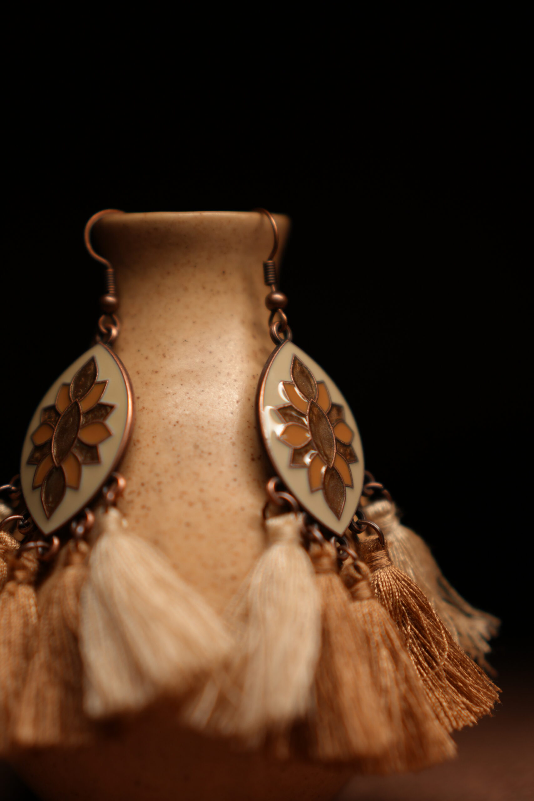 A pair of ivory and gold lotus enamel marquise-shaped drop earrings with five alternating ivory cream and champagne gold thread tassels, displayed on a speckled beige ceramic vase against a dark background.