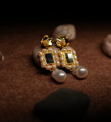 A pair of gold-tone drop earrings with a sculpted floral stud top, a rectangular kundan-style mirror centre bordered by faux pearls, and a baroque pearl drop, displayed on smooth pebbles against a warm brown fabric background.