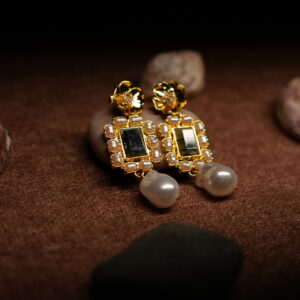 A pair of gold-tone drop earrings with a sculpted floral stud top, a rectangular kundan-style mirror centre bordered by faux pearls, and a baroque pearl drop, displayed on smooth pebbles against a warm brown fabric background.