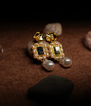 A pair of gold-tone drop earrings with a sculpted floral stud top, a rectangular kundan-style mirror centre bordered by faux pearls, and a baroque pearl drop, displayed on smooth pebbles against a warm brown fabric background.