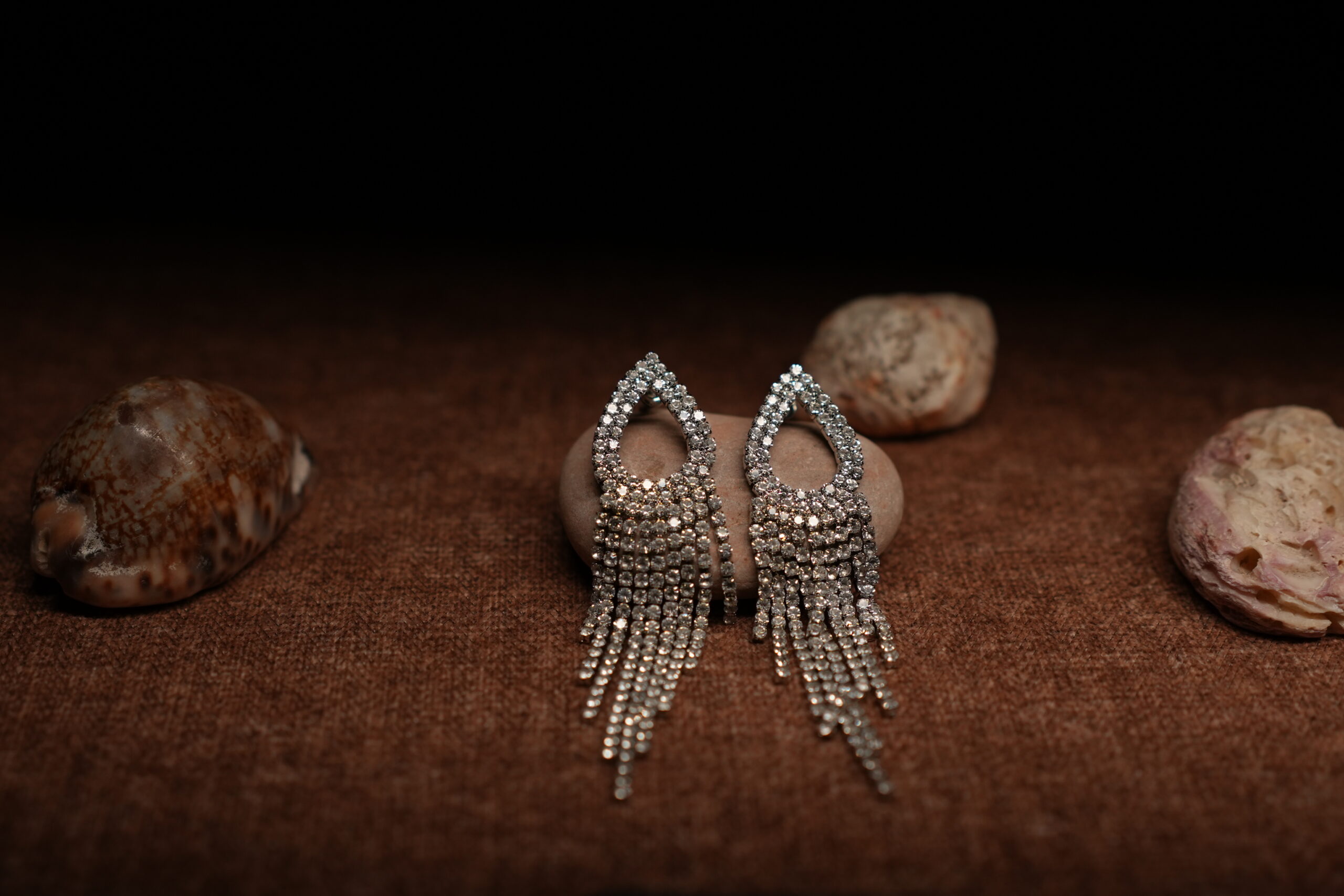 A pair of silver-tone fully pave crystal open teardrop earrings with eight graduated rhinestone chain fringe strands, displayed on a smooth beige pebble surrounded by seashells against a warm dark background.