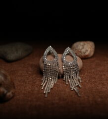 A pair of silver-tone fully pave crystal open teardrop earrings with eight graduated rhinestone chain fringe strands, displayed on a smooth beige pebble surrounded by seashells against a warm dark background.