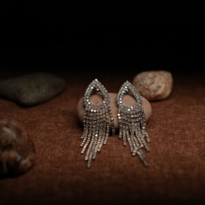A pair of silver-tone fully pave crystal open teardrop earrings with eight graduated rhinestone chain fringe strands, displayed on a smooth beige pebble surrounded by seashells against a warm dark background.