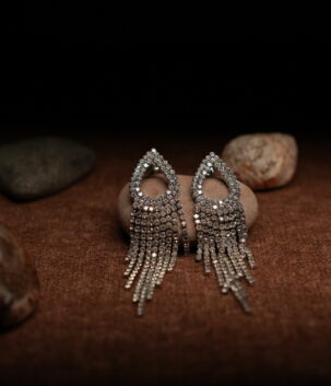 A pair of silver-tone fully pave crystal open teardrop earrings with eight graduated rhinestone chain fringe strands, displayed on a smooth beige pebble surrounded by seashells against a warm dark background.