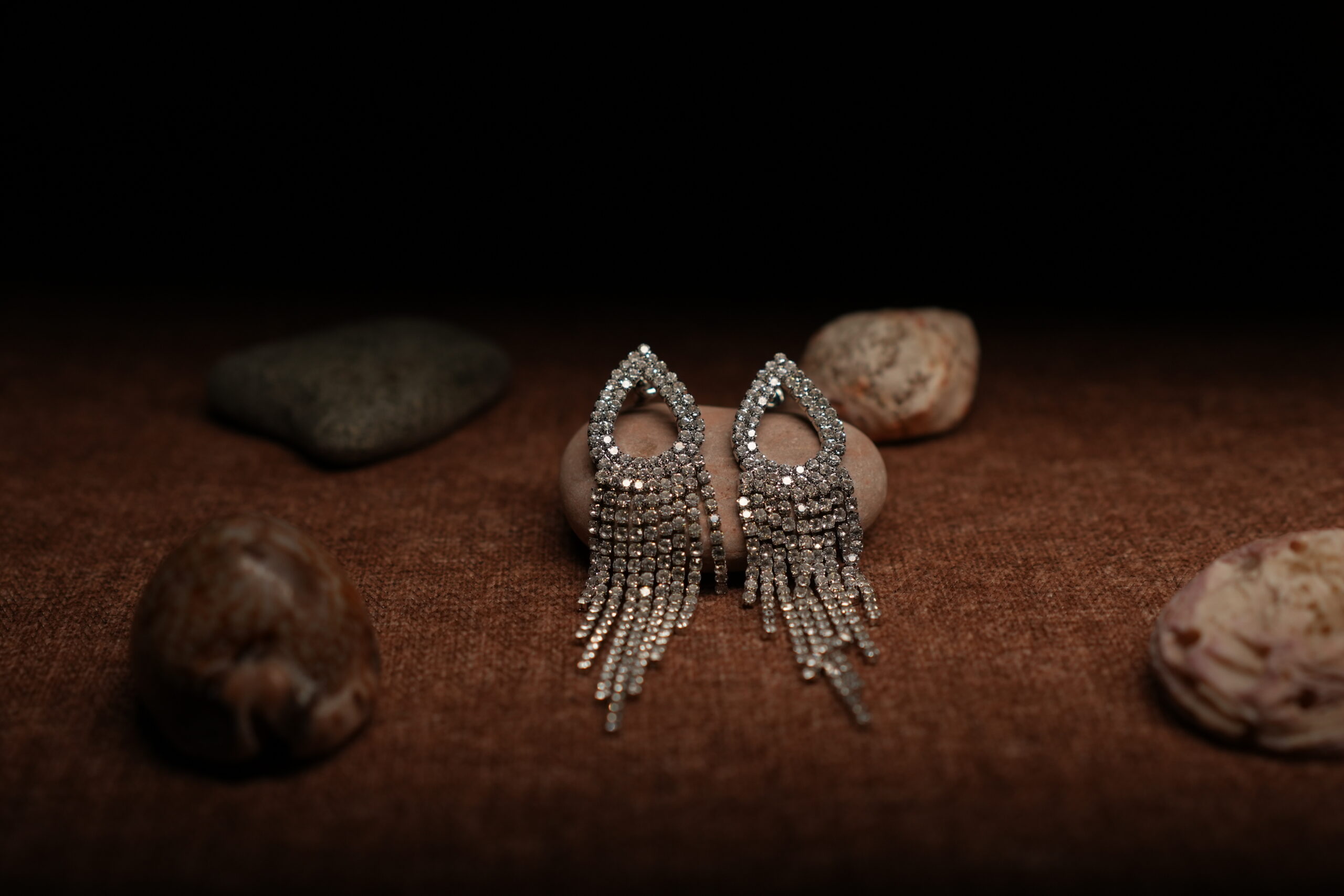 A pair of silver-tone fully pave crystal open teardrop earrings with eight graduated rhinestone chain fringe strands, displayed on a smooth beige pebble surrounded by seashells against a warm dark background.