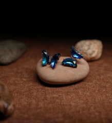 A pair of gold-tone butterfly stud earrings assembled from faceted ocean blue and teal marquise crystals in a full wing silhouette, displayed on a smooth beige pebble against a dark background.
