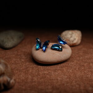A pair of gold-tone butterfly stud earrings assembled from faceted ocean blue and teal marquise crystals in a full wing silhouette, displayed on a smooth beige pebble against a dark background.