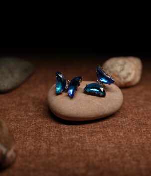 A pair of gold-tone butterfly stud earrings assembled from faceted ocean blue and teal marquise crystals in a full wing silhouette, displayed on a smooth beige pebble against a dark background.