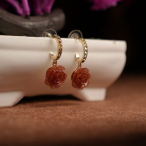 A pair of gold-tone pave crystal hoop earrings with a sculpted amber glitter resin rose pendant, displayed on a white ceramic planter with purple dried flowers in the background against a dark setting.