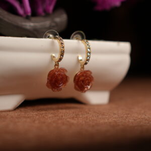 A pair of gold-tone pave crystal hoop earrings with a sculpted amber glitter resin rose pendant, displayed on a white ceramic planter with purple dried flowers in the background against a dark setting.