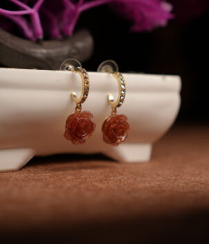 A pair of gold-tone pave crystal hoop earrings with a sculpted amber glitter resin rose pendant, displayed on a white ceramic planter with purple dried flowers in the background against a dark setting.