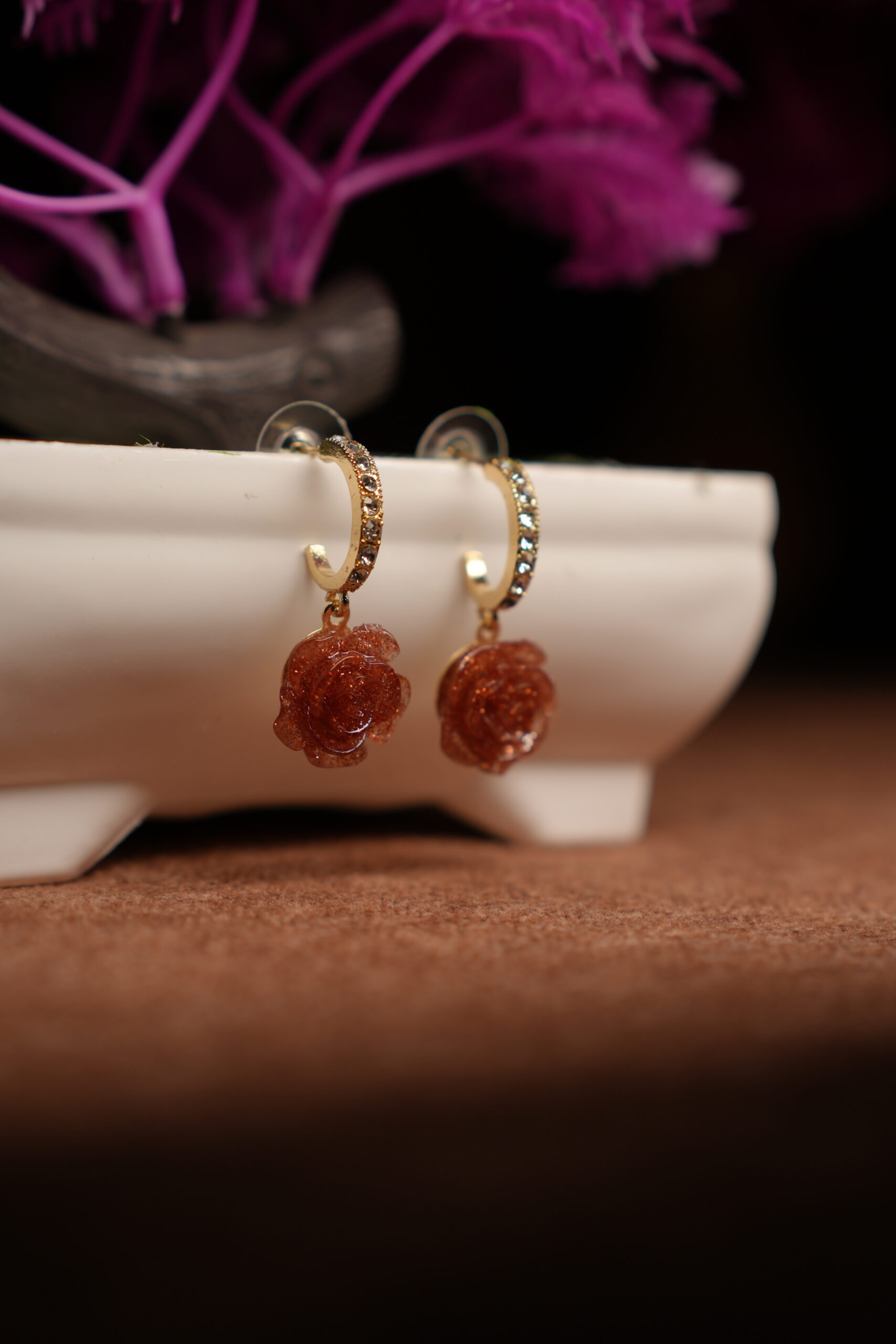 A pair of gold-tone pave crystal hoop earrings with a sculpted amber glitter resin rose pendant, displayed on a white ceramic planter with purple dried flowers in the background against a dark setting.