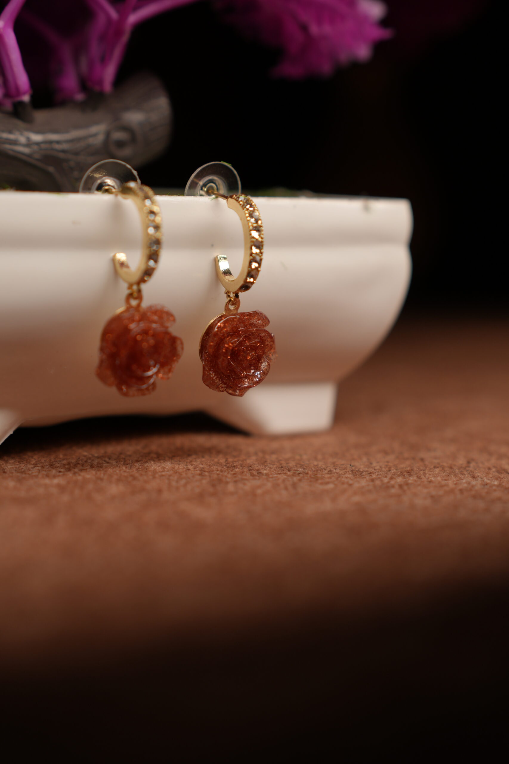 A pair of gold-tone pave crystal hoop earrings with a sculpted amber glitter resin rose pendant, displayed on a white ceramic planter with purple dried flowers in the background against a dark setting.