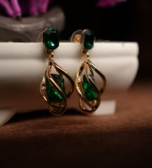 A pair of gold-tone drop earrings with a faceted emerald green square crystal stud top and an open twist cage pendant enclosing a matching emerald green teardrop crystal, displayed on a white ceramic planter with purple dried flowers in the background.