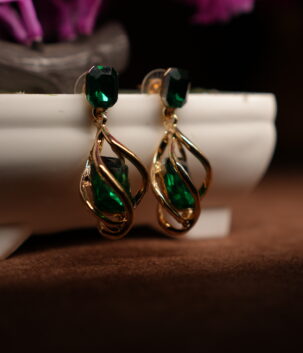A pair of gold-tone drop earrings with a faceted emerald green square crystal stud top and an open twist cage pendant enclosing a matching emerald green teardrop crystal, displayed on a white ceramic planter with purple dried flowers in the background.