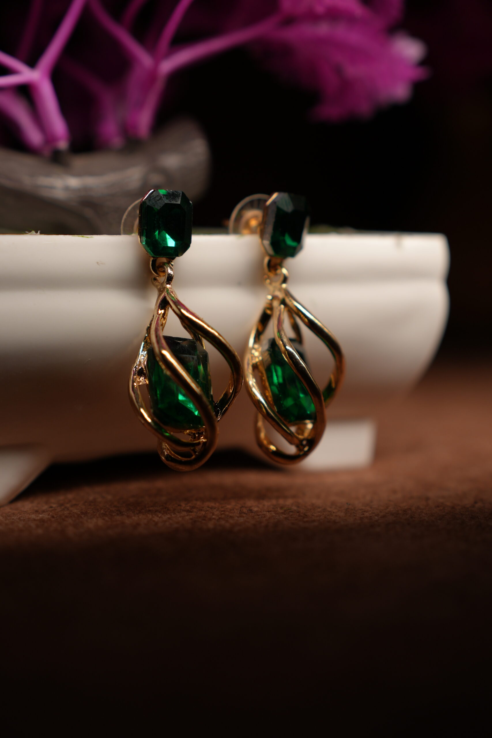 A pair of gold-tone drop earrings with a faceted emerald green square crystal stud top and an open twist cage pendant enclosing a matching emerald green teardrop crystal, displayed on a white ceramic planter with purple dried flowers in the background.
