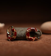 A pair of gold-tone hoop earrings with deep crimson red glossy enamel fill and scalloped gold edge detailing, resting against a dark grey stone on a warm brown fabric surface.