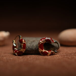 A pair of gold-tone hoop earrings with deep crimson red glossy enamel fill and scalloped gold edge detailing, resting against a dark grey stone on a warm brown fabric surface.