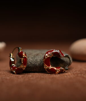 A pair of gold-tone hoop earrings with deep crimson red glossy enamel fill and scalloped gold edge detailing, resting against a dark grey stone on a warm brown fabric surface.