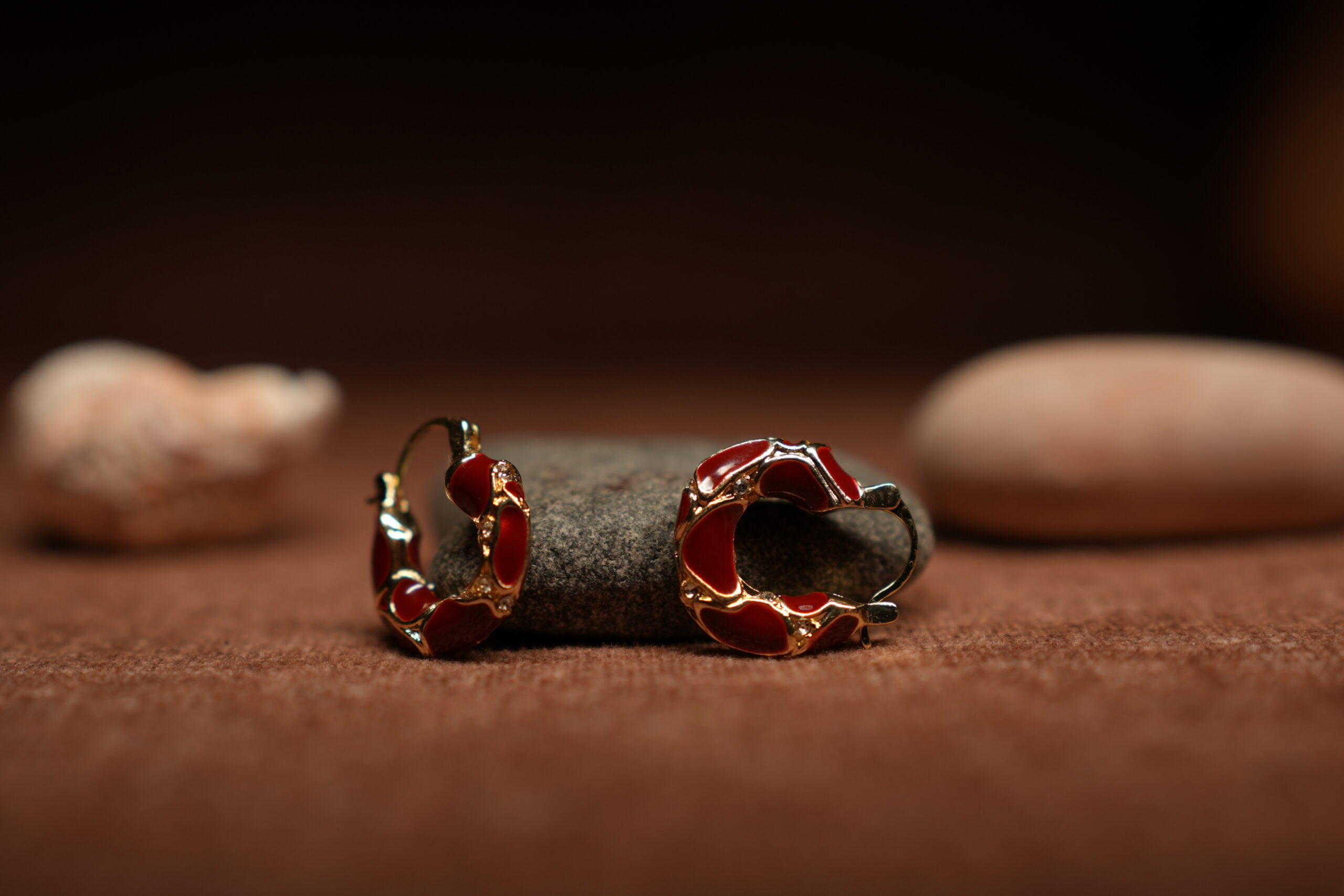 A pair of gold-tone hoop earrings with deep crimson red glossy enamel fill and scalloped gold edge detailing, resting against a dark grey stone on a warm brown fabric surface.