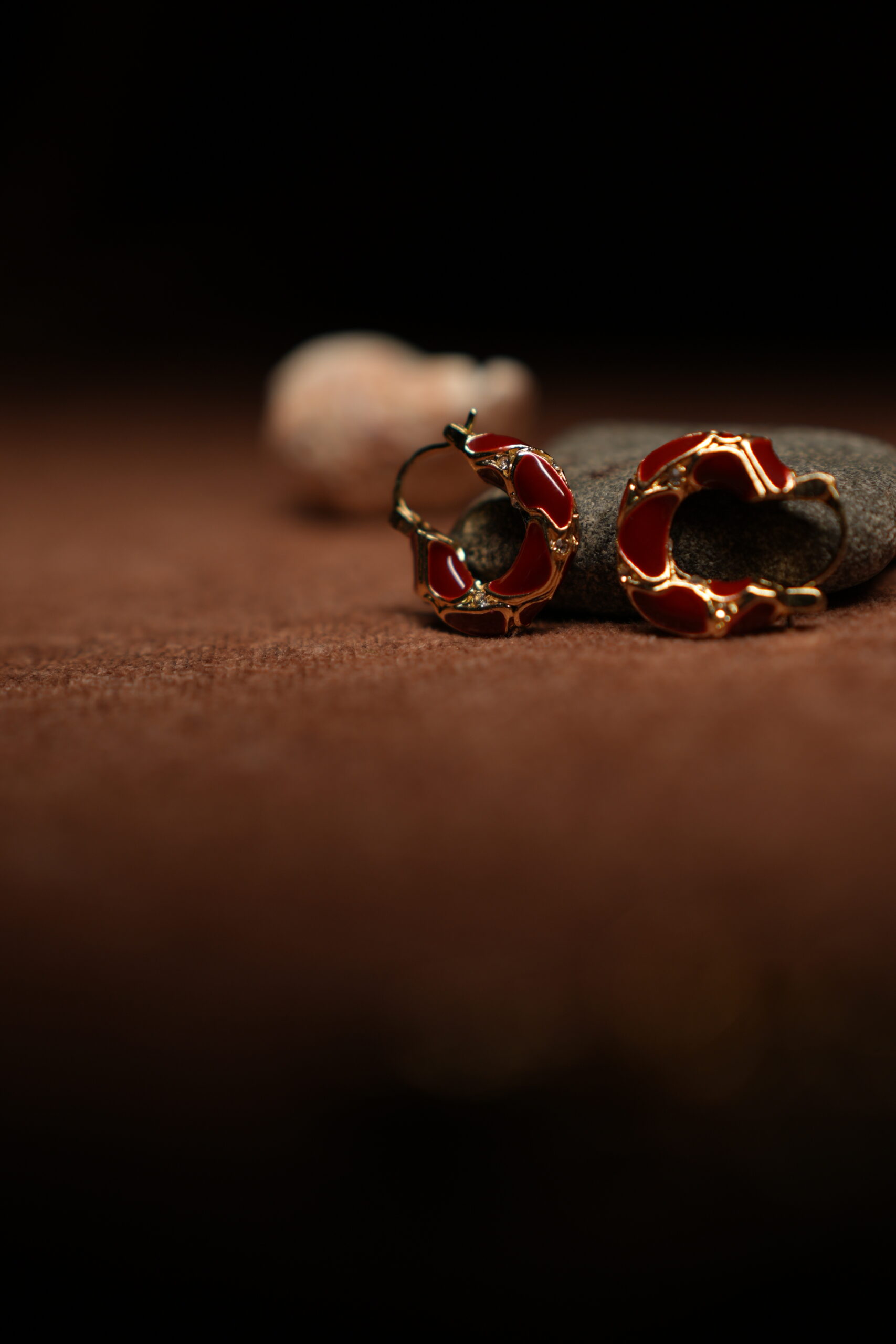 A pair of gold-tone hoop earrings with deep crimson red glossy enamel fill and scalloped gold edge detailing, resting against a dark grey stone on a warm brown fabric surface.