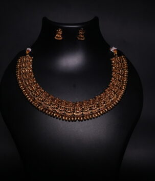 Antique gold plated temple jewellery set featuring an intricately engraved goddess motif necklace with matching jhumka earrings, displayed on a black velvet bust.