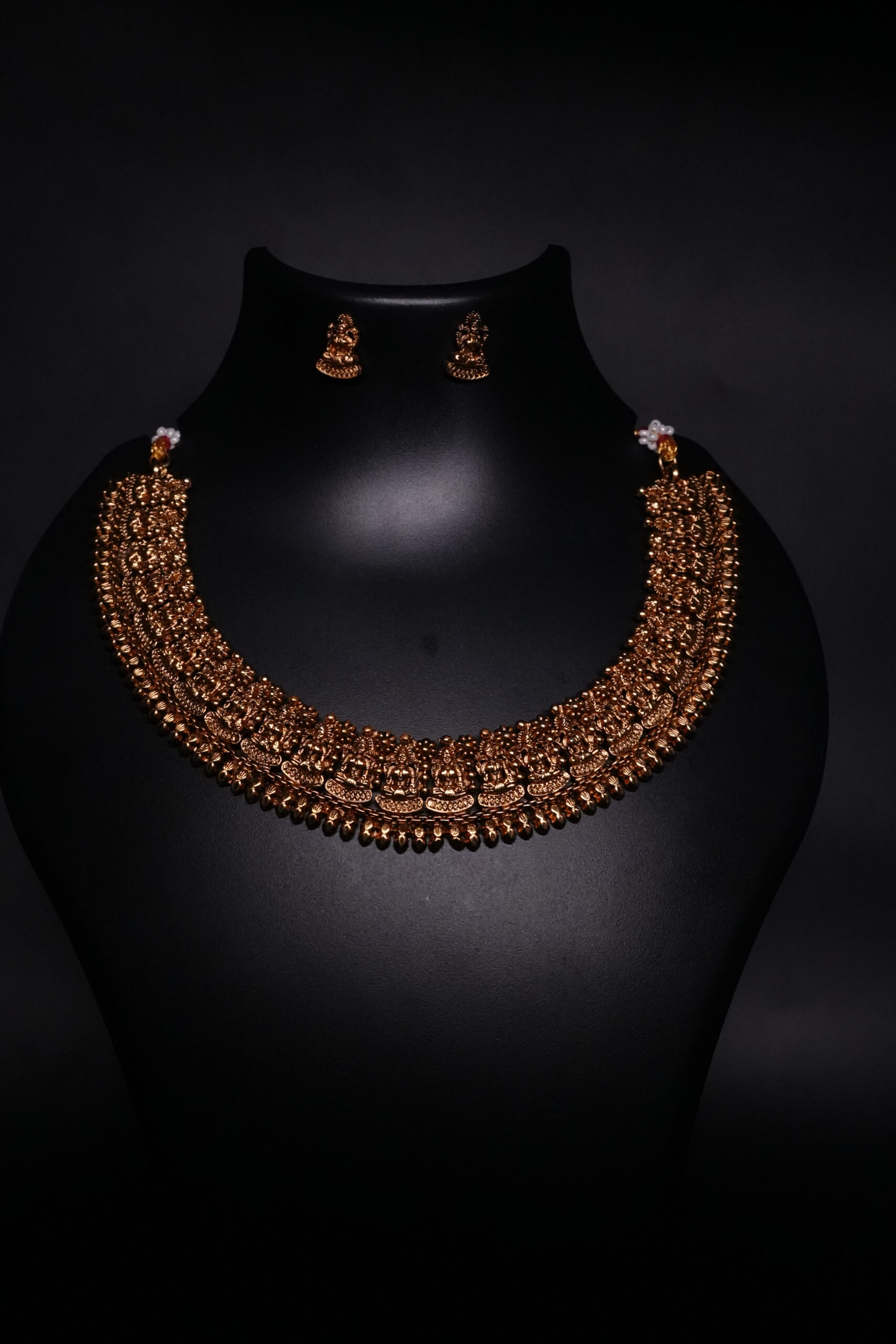 Antique gold plated temple jewellery set featuring an intricately engraved goddess motif necklace with matching jhumka earrings, displayed on a black velvet bust.
