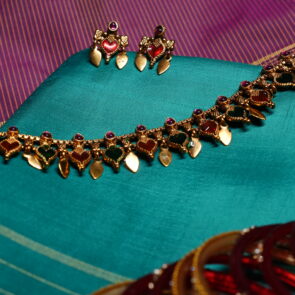 Antique gold plated necklace and earring set with red and green kemp stone heart motifs, displayed on a teal silk saree.