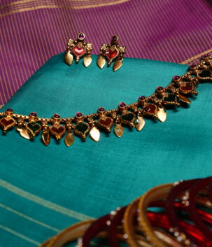 Antique gold plated necklace and earring set with red and green kemp stone heart motifs, displayed on a teal silk saree.