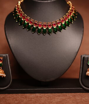 Gold plated meenakari choker necklace with green and red enamel leaf motifs, paired with matching pearl jhumka earrings on a black display bust.