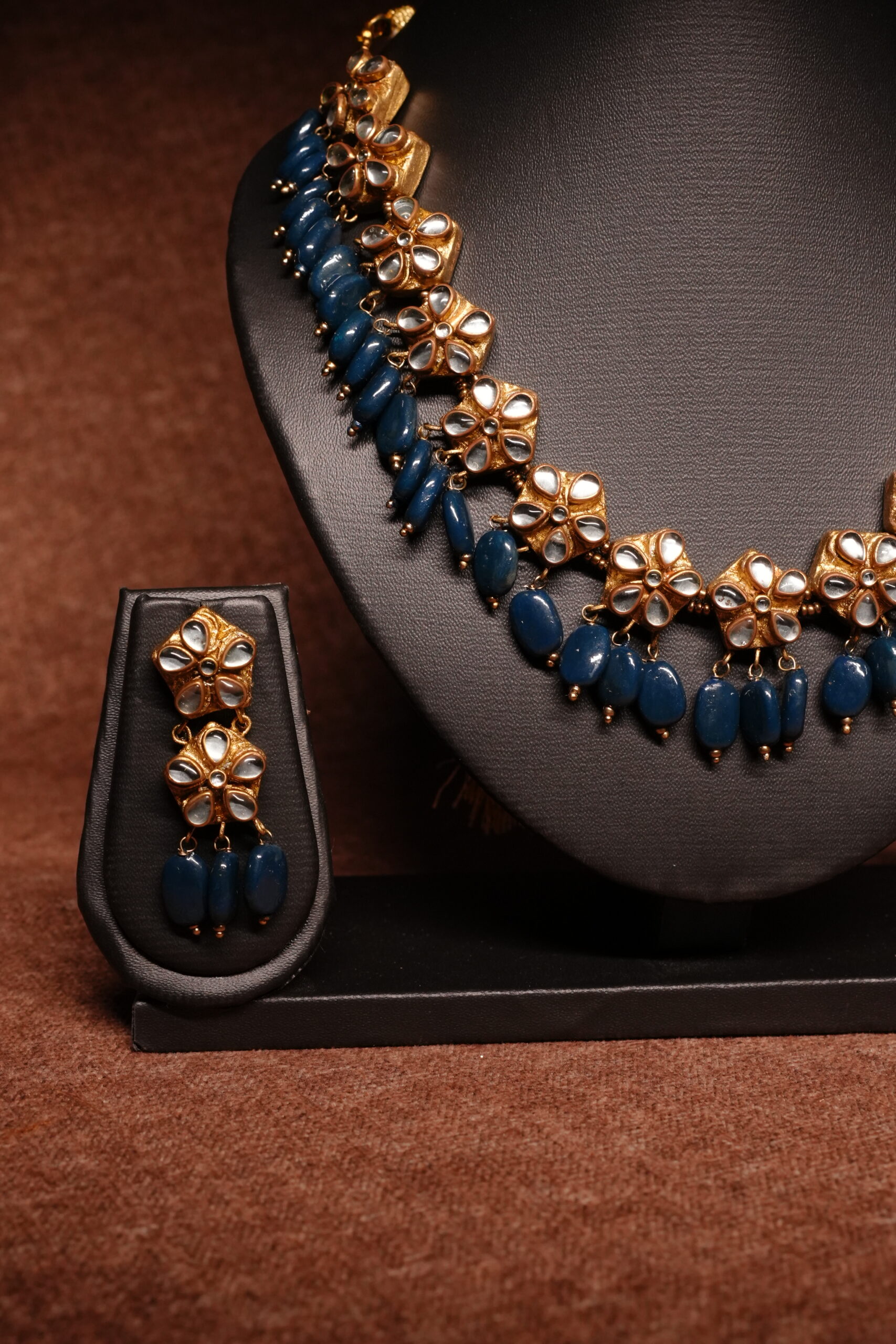 Gold plated kundan necklace set with floral motifs and deep blue beaded drops, paired with matching earrings on a black display bus