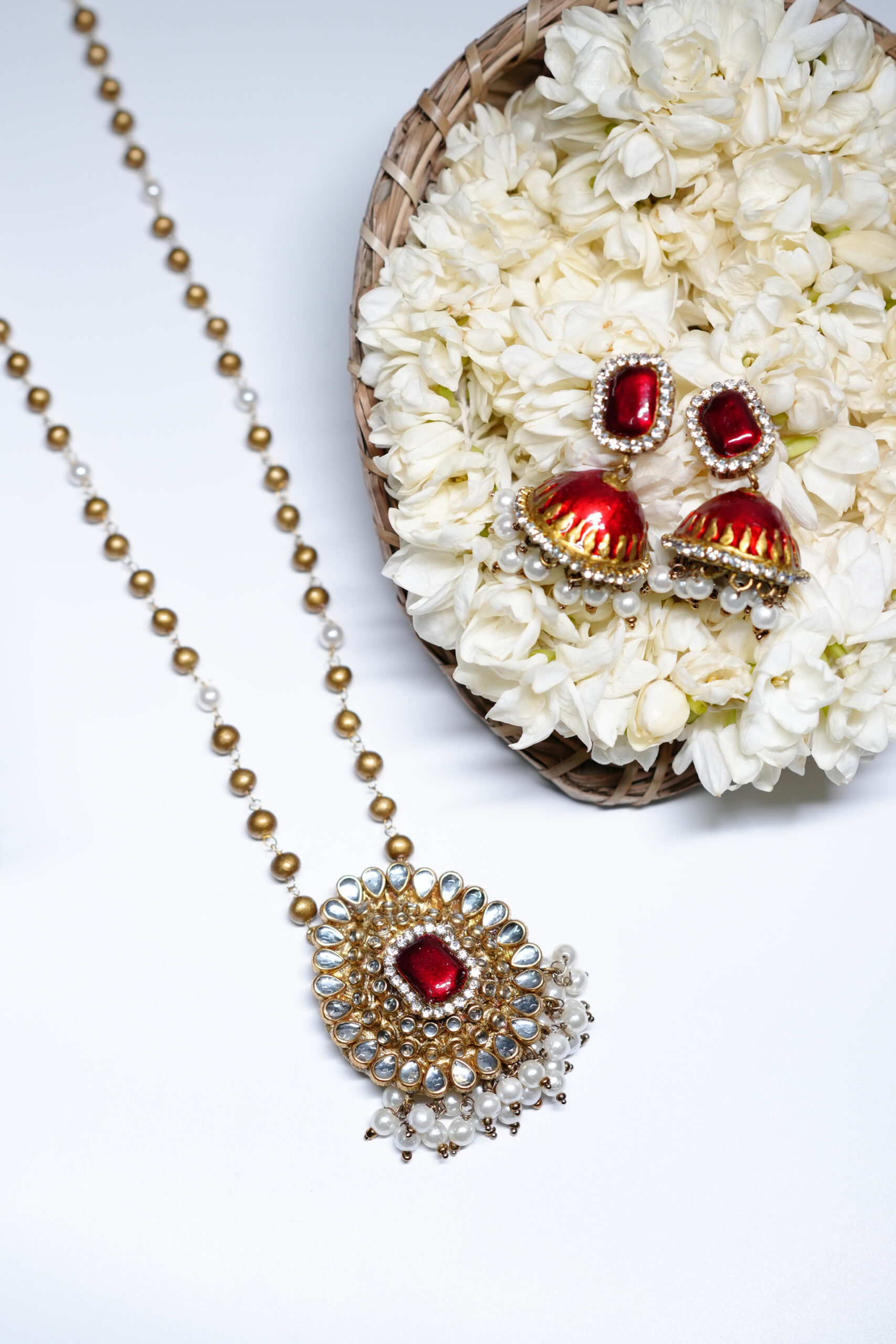Gold plated kundan pendant necklace set with red stone and pearl jhumka earrings placed on white jasmine flowers.
