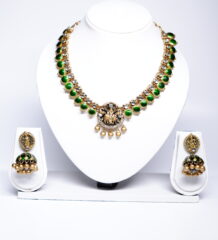 Gold plated temple-style necklace and jhumka earring set with green kundan stones and a deity motif, displayed on a white jewellery stand.