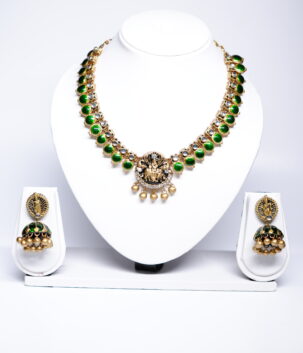 Gold plated temple-style necklace and jhumka earring set with green kundan stones and a deity motif, displayed on a white jewellery stand.