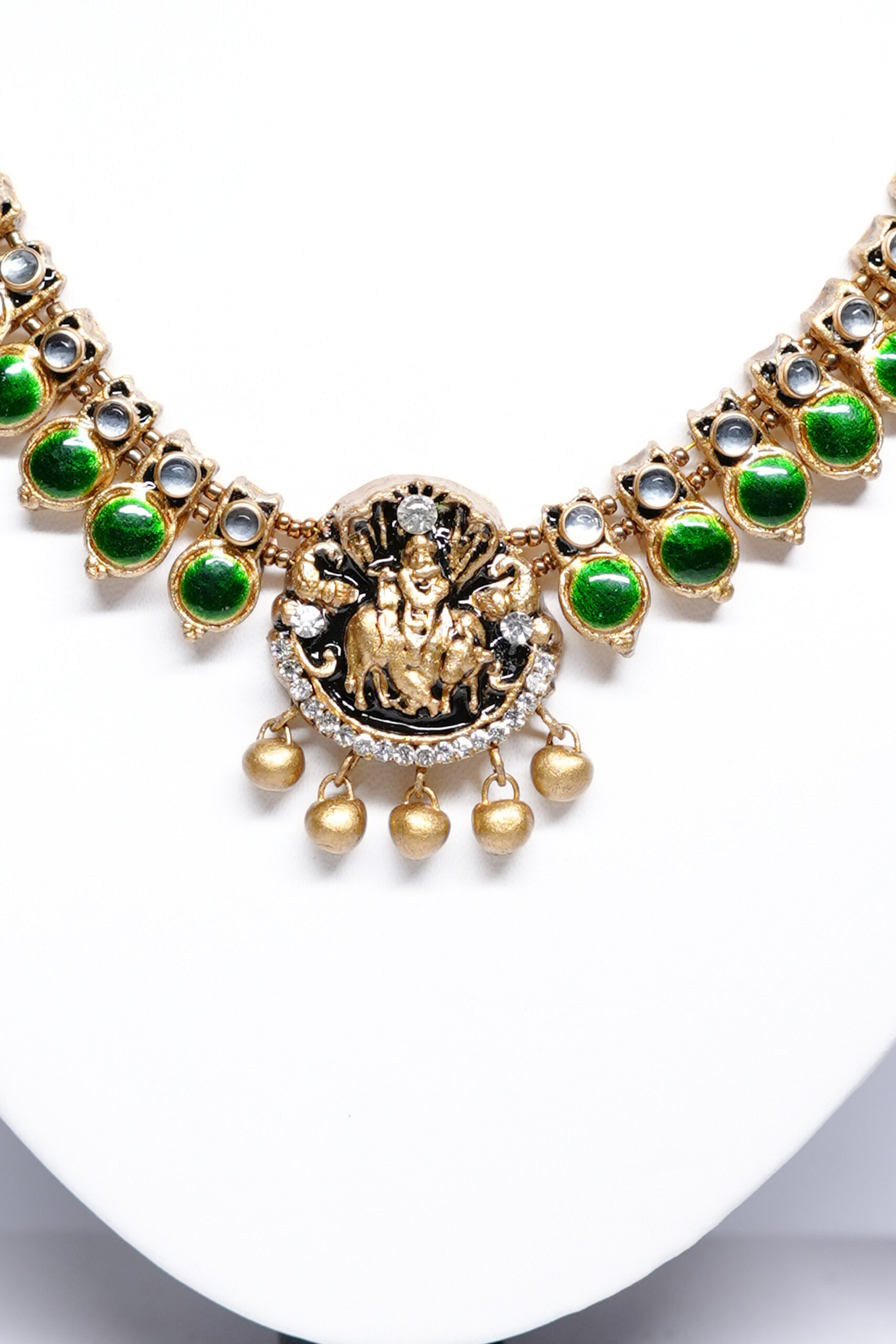 Gold plated kundan necklace set with green stones and temple-style deity pendant displayed on a jewellery stand.