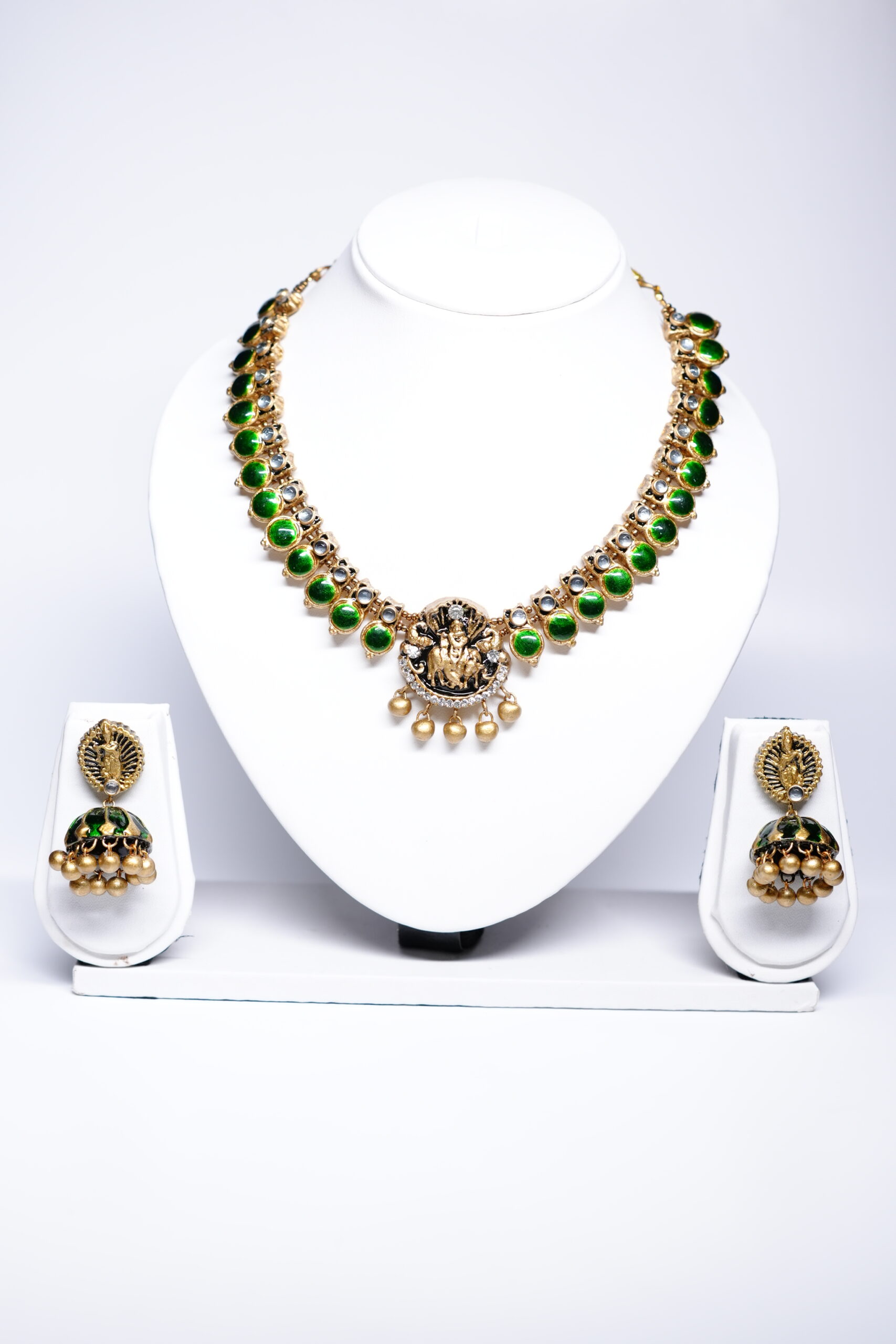 Gold plated temple-style necklace and jhumka earring set with green kundan stones and a deity motif, displayed on a white jewellery stand.