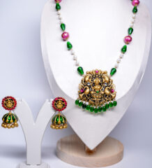 Antique gold plated temple-style necklace set with a goddess Lakshmi pendant, emerald-green beads, pink meenakari accents, and matching jhumka earrings, displayed on a white velvet bust.