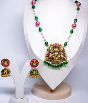 Antique gold plated temple-style necklace set with a goddess Lakshmi pendant, emerald-green beads, pink meenakari accents, and matching jhumka earrings, displayed on a white velvet bust.