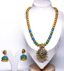 Gold plated beaded necklace with blue and green meenakari peacock pendant and matching jhumka earrings displayed on a jewellery stand.
