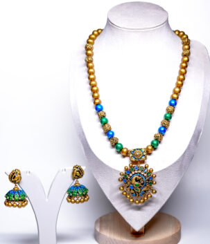 Gold plated beaded necklace with blue and green meenakari peacock pendant and matching jhumka earrings displayed on a jewellery stand.