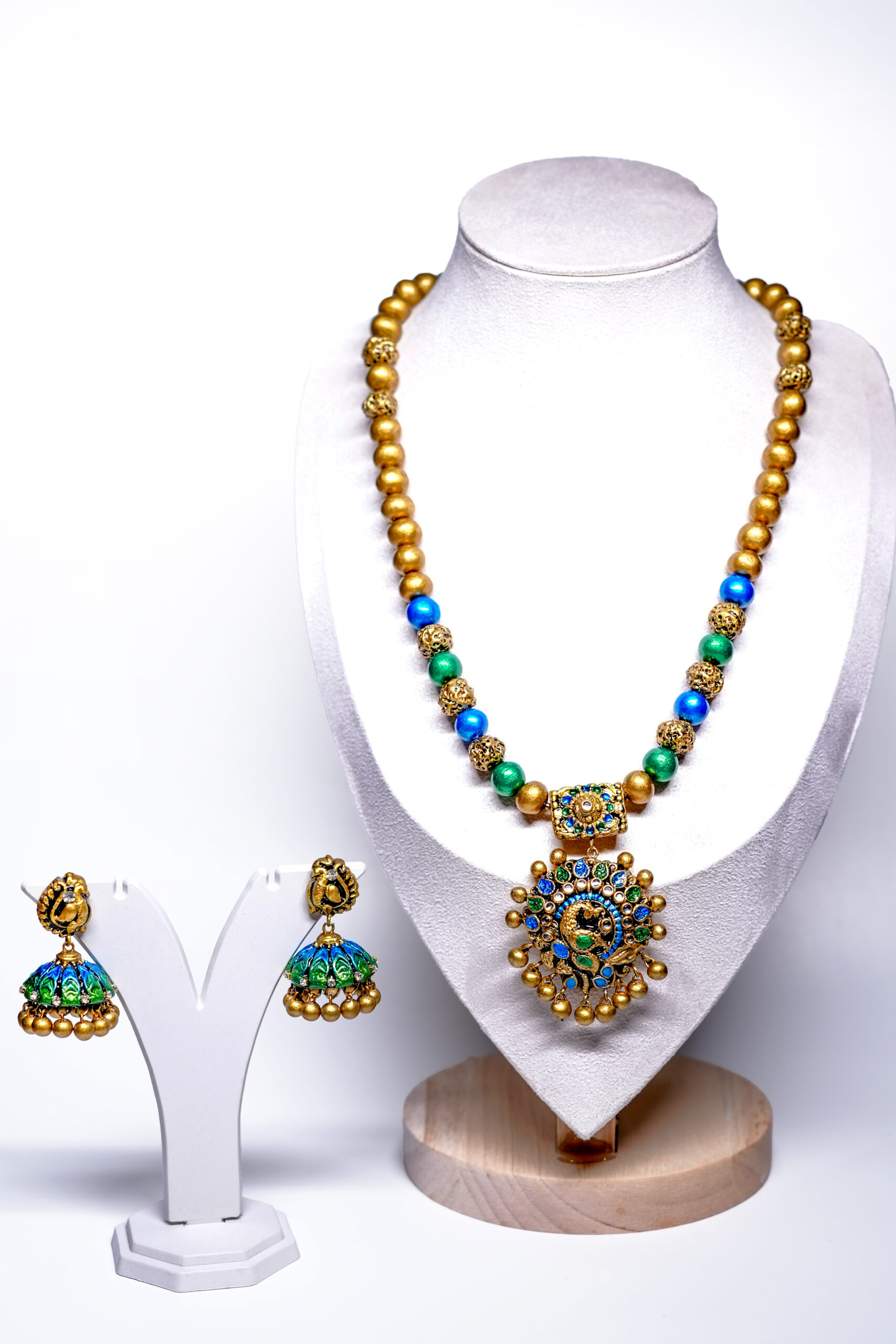 Gold plated beaded necklace with blue and green meenakari peacock pendant and matching jhumka earrings displayed on a jewellery stand.
