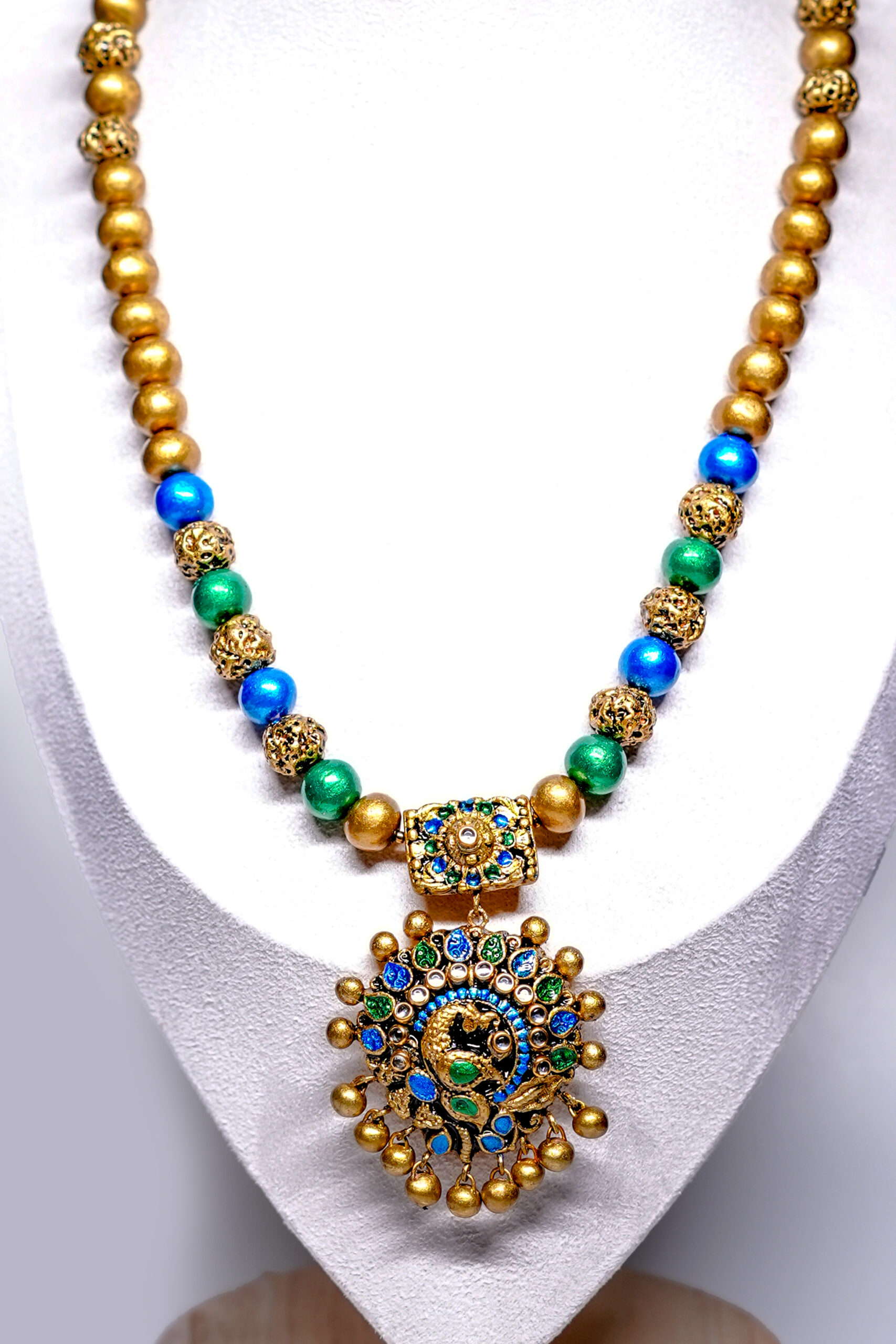 Gold plated beaded necklace with blue and green meenakari peacock pendant displayed on a white jewellery bust, with matching jhumka earrings alongside.