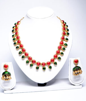 Gold plated necklace with alternating red and green kemp stones and matching jhumka earrings displayed on a white jewellery bust.